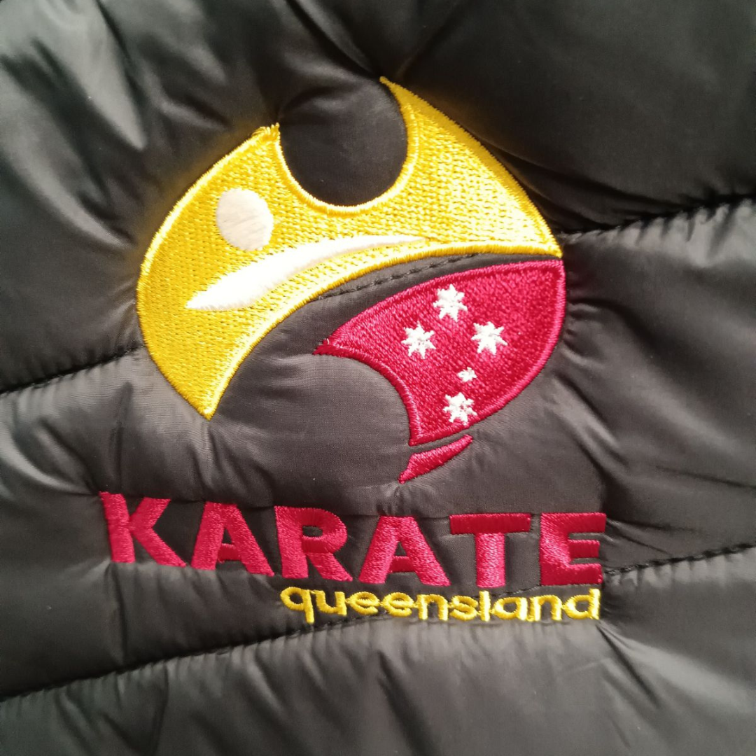 Detailed embroidery of the Karate Queensland logo on the chest of a black puffer jacket.